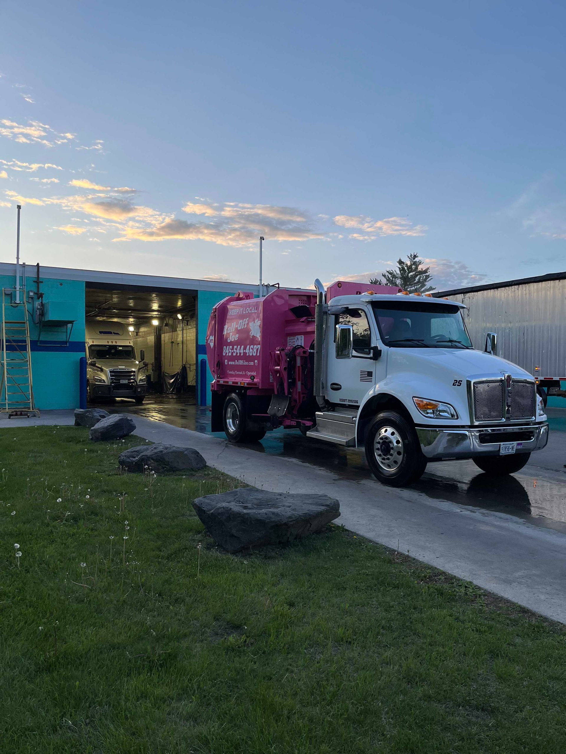 White and pink garbage truck exiting a teal building with a car wash, under a cloudy sky.