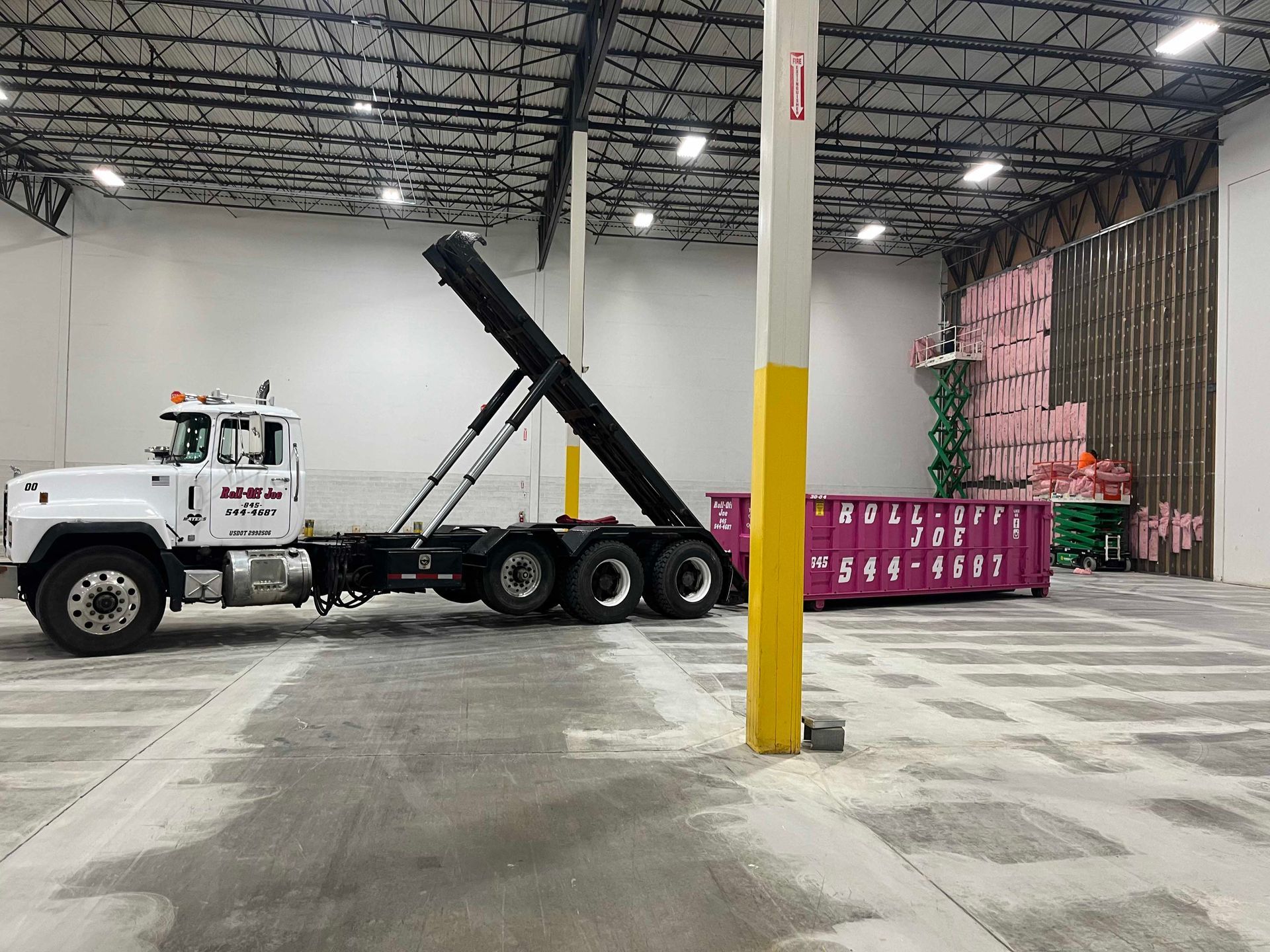 White truck in a warehouse with a dumpster. The truck's arm is raised, next to a pink dumpster.