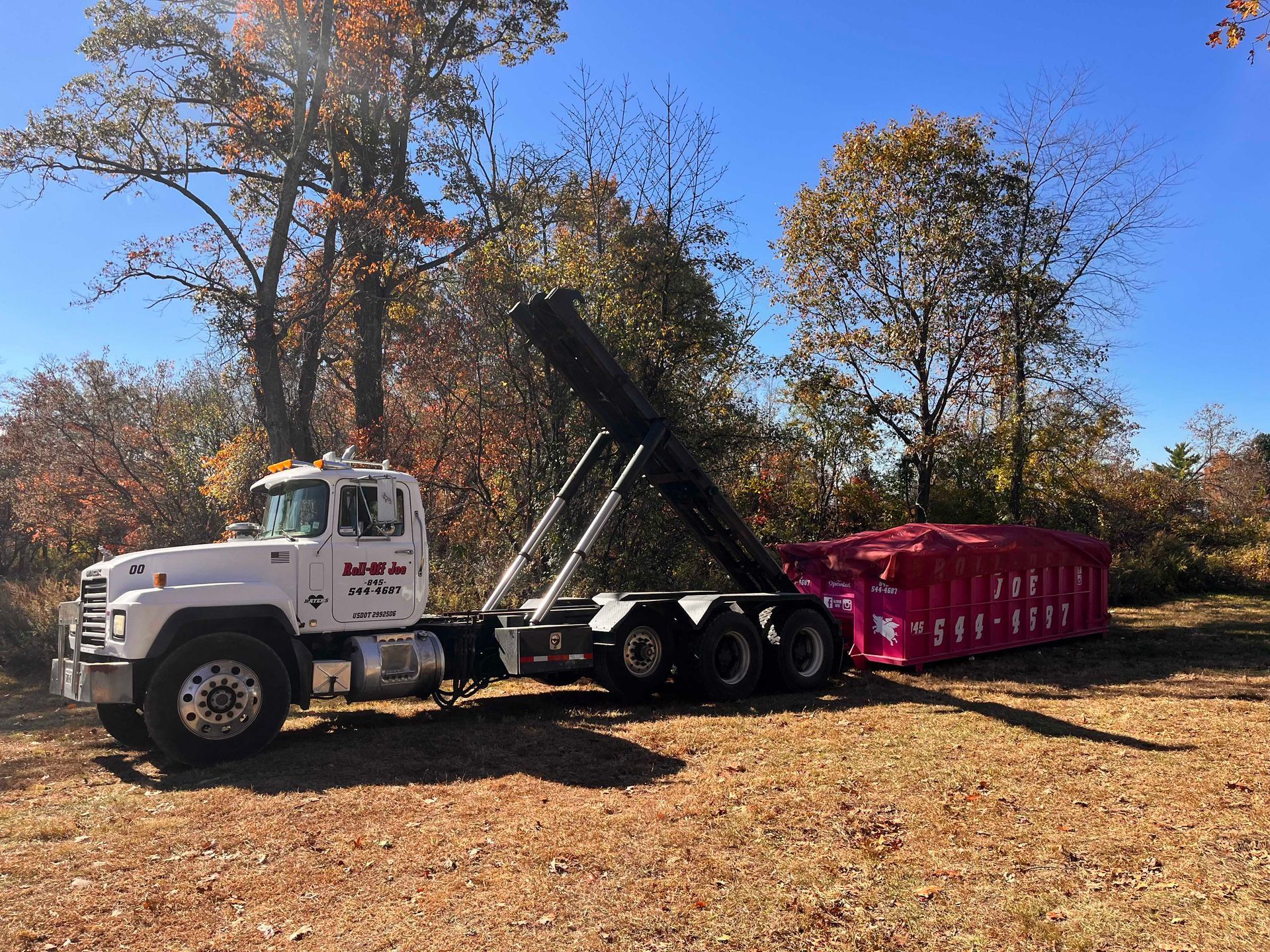 White truck with lift attached to a pink container in a field with trees in the background.
