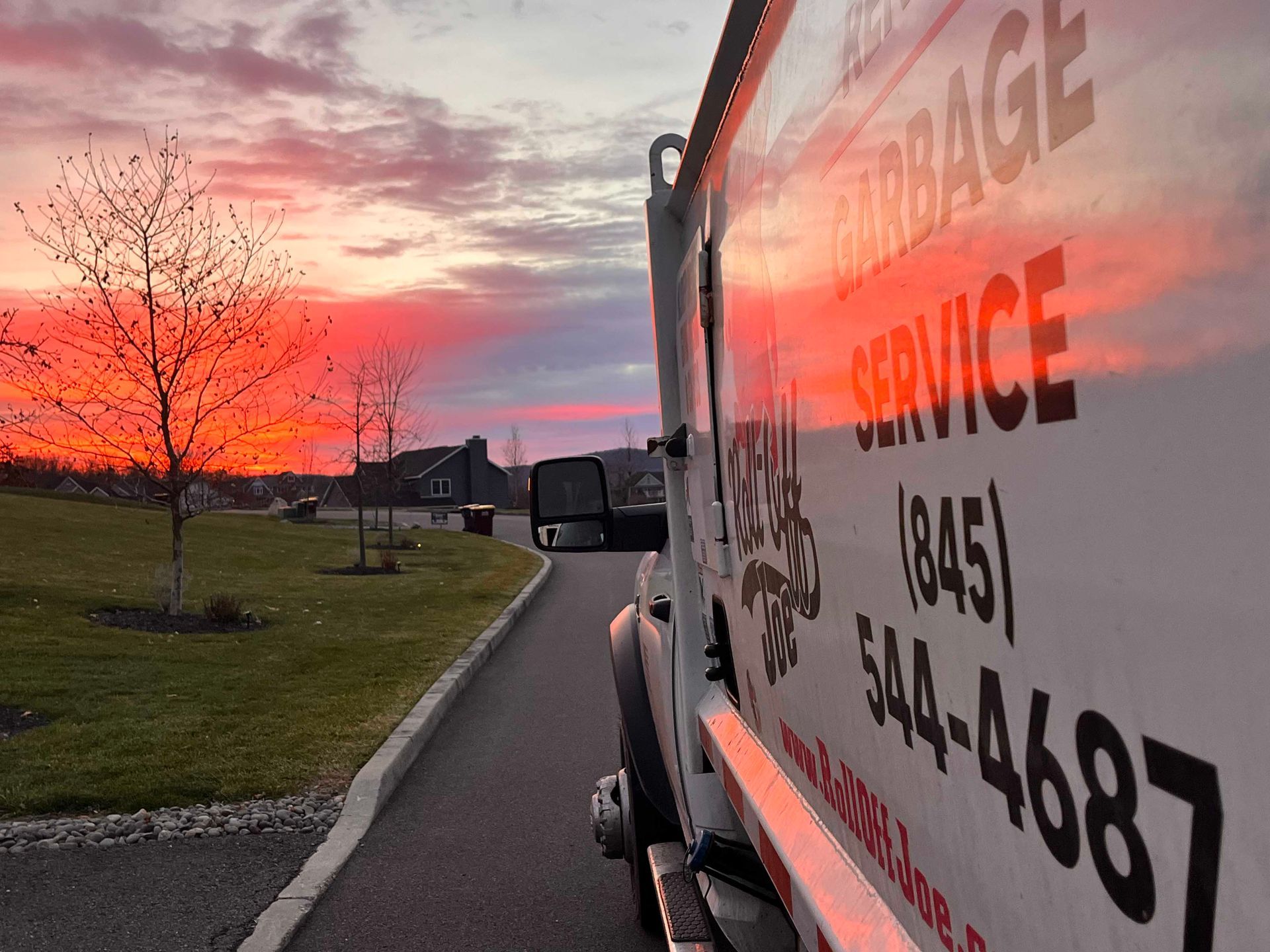 Garbage truck with service details on the side, parked at dusk with a vibrant, orange and pink sunset in the background.