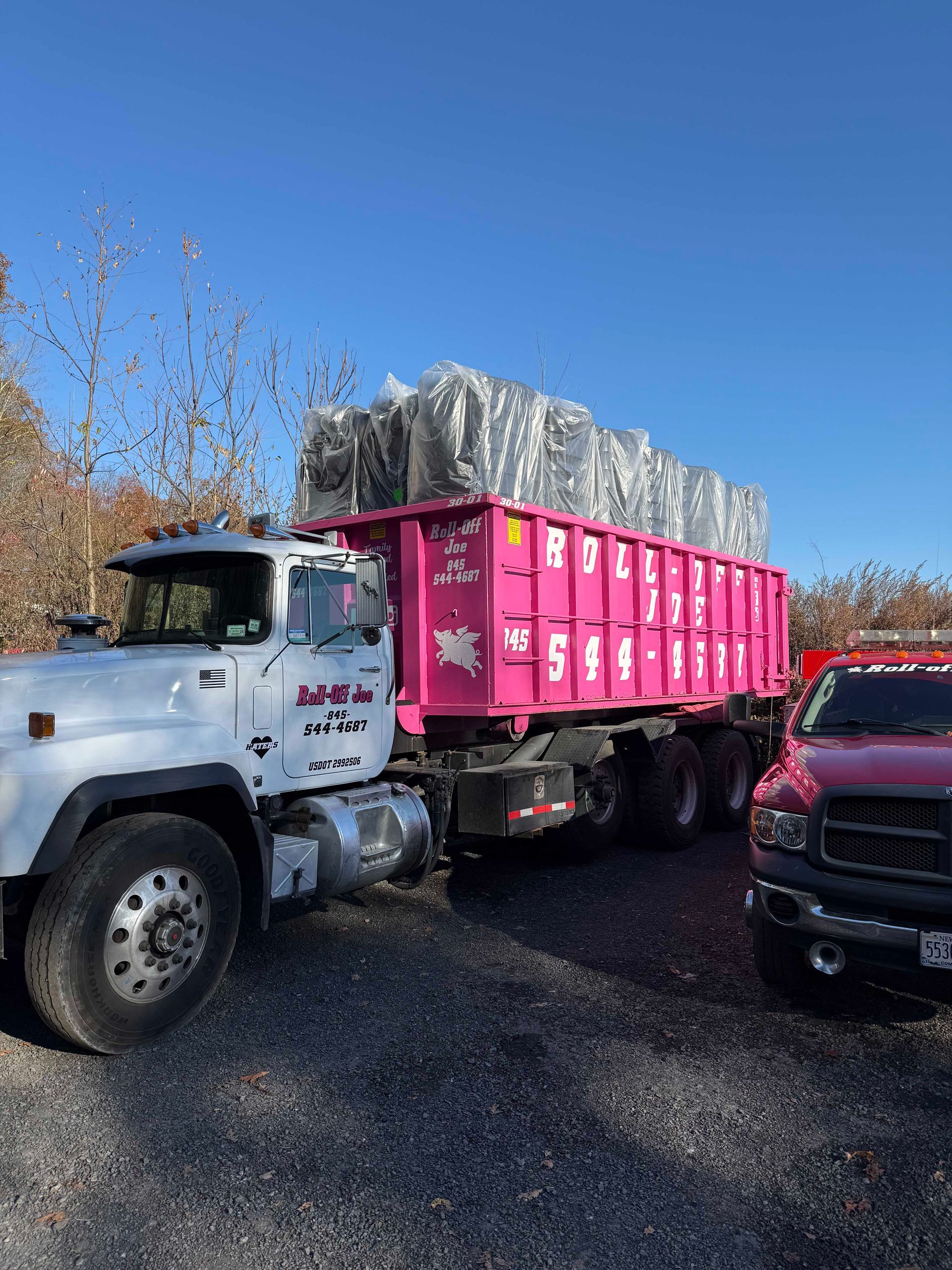 White truck with pink dumpster filled with debris; red truck parked to the side. Clear blue sky.