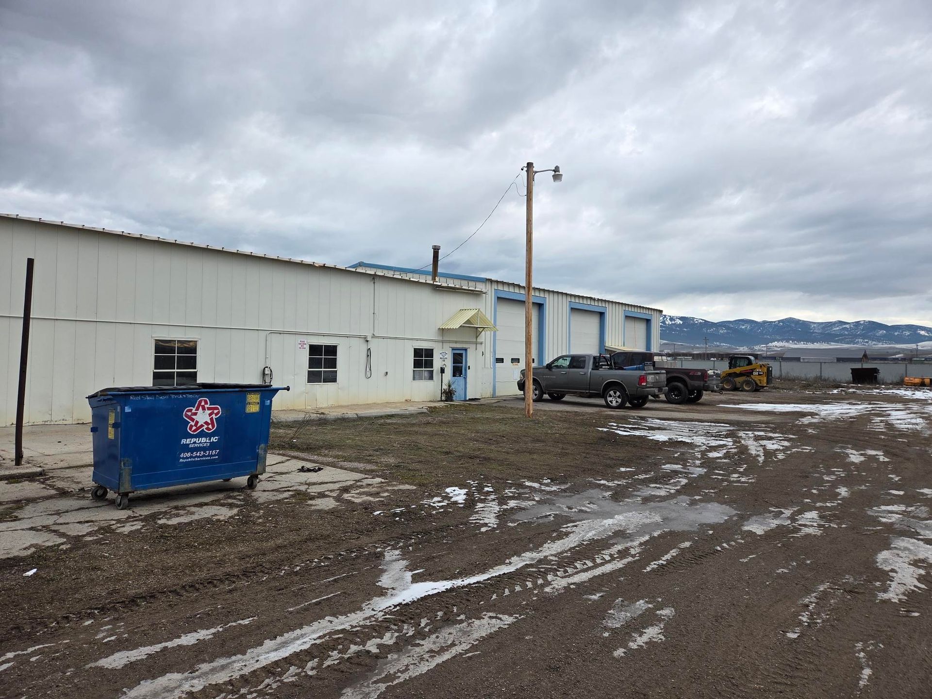 Exterior of a white industrial building with a dumpster and muddy ground. A truck and a backhoe are parked nearby.