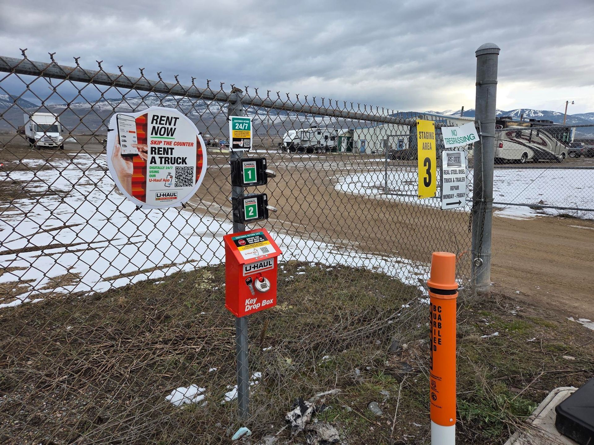 Chain-link fence with signs attached, including a red box. An orange post is on the right. Cloudy sky.