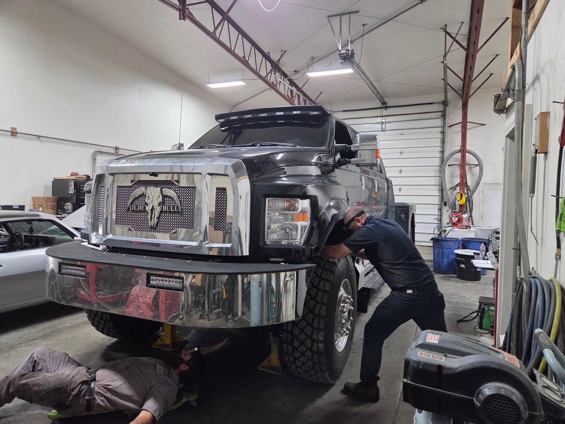 Men working on a large black truck in a garage; one under the truck, the other by the front tire.