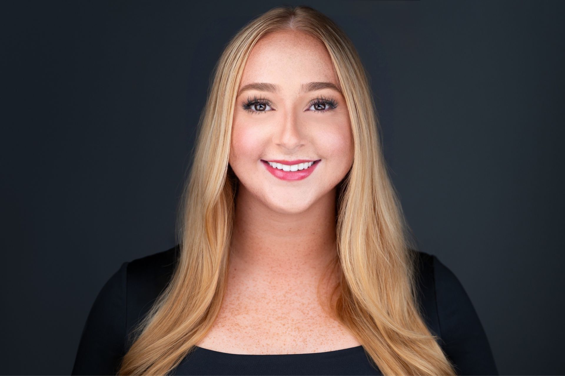 Blond woman with a bright smile, wearing a black top, against a dark background. Rebecca Saitta dental assistant and front desk receptionist