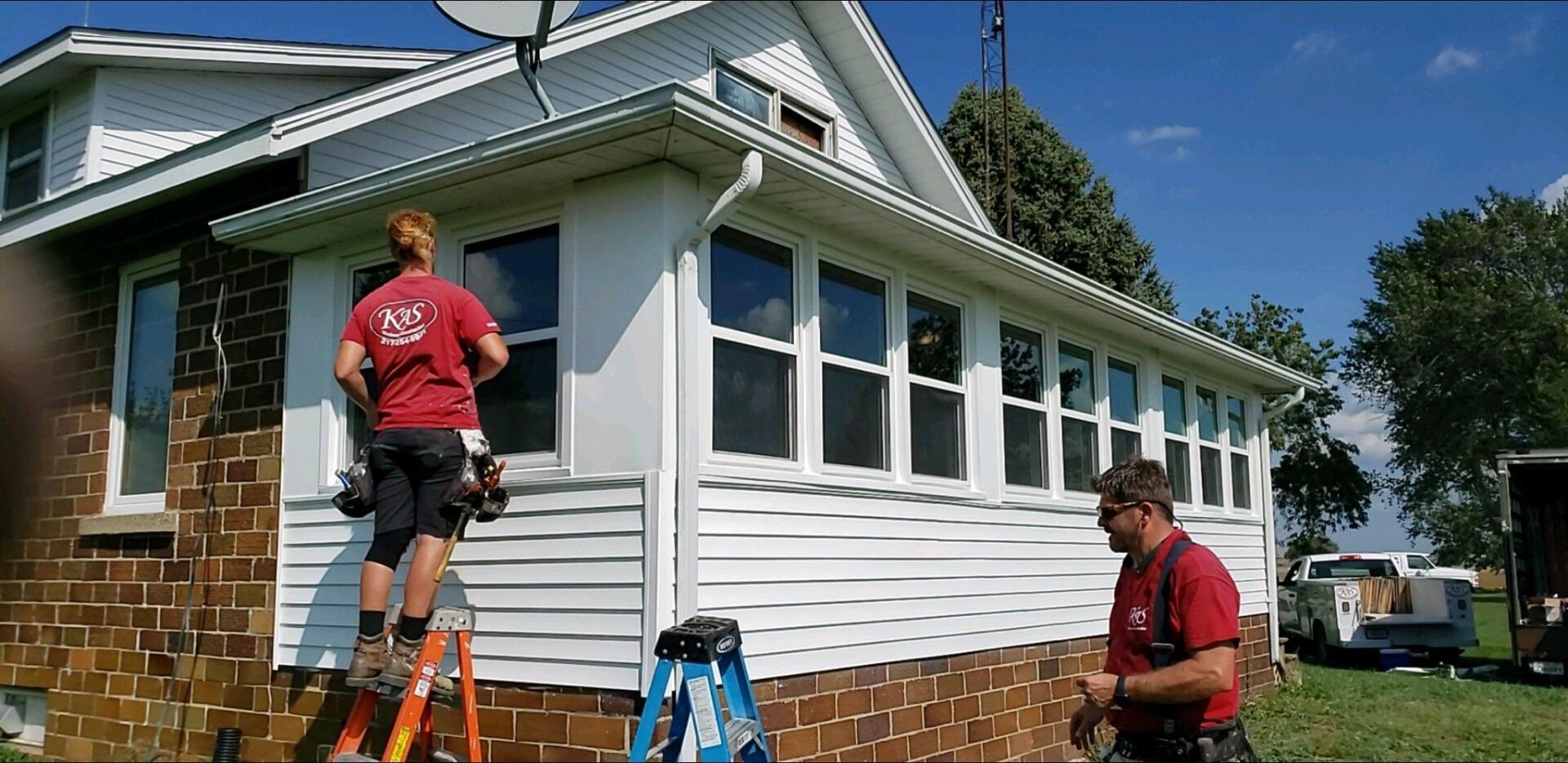 Two men installing windows on a white house with red shirts on a sunny day.