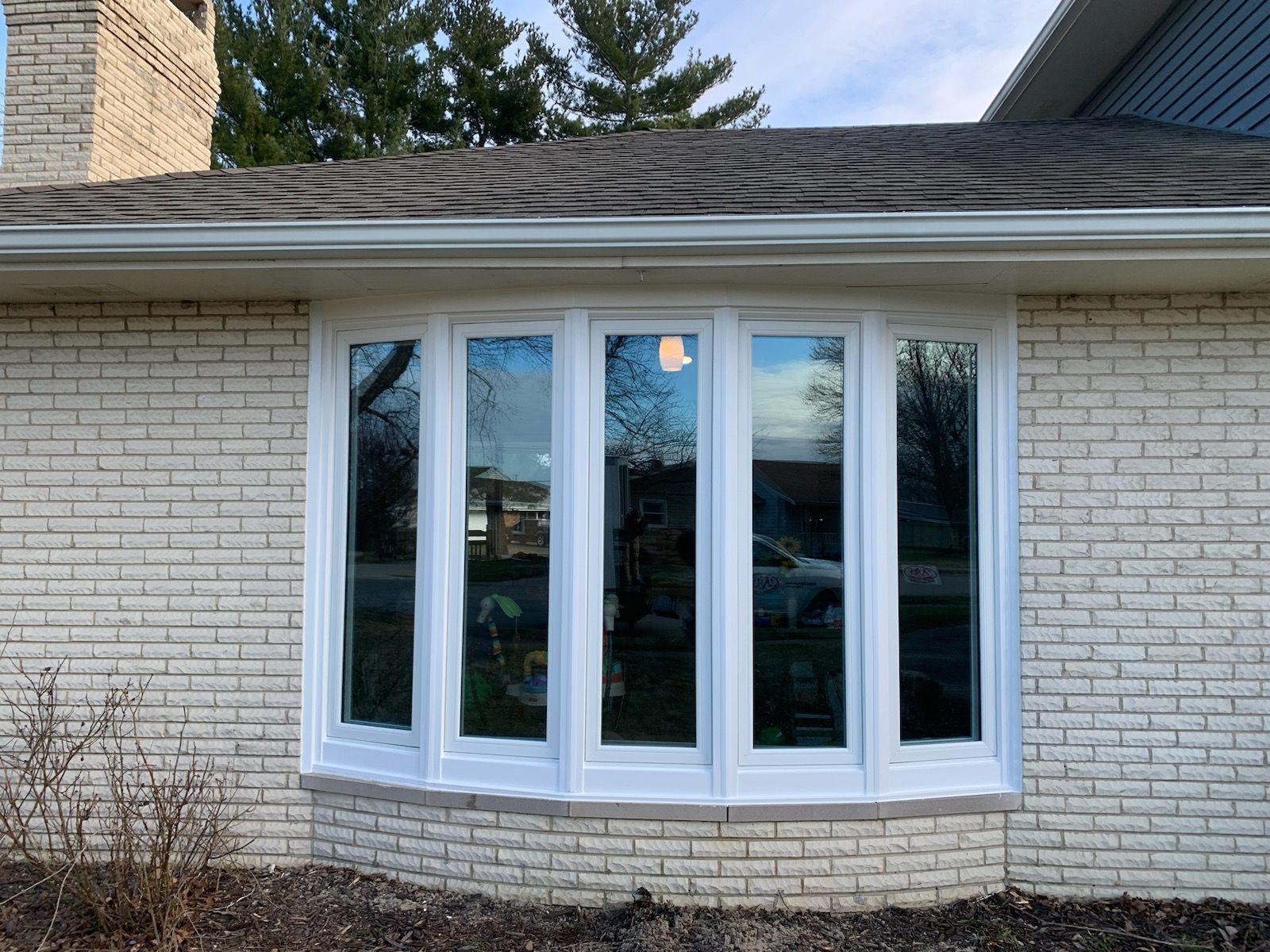 White bay window in a brick house. Reflected trees and sky in the glass.