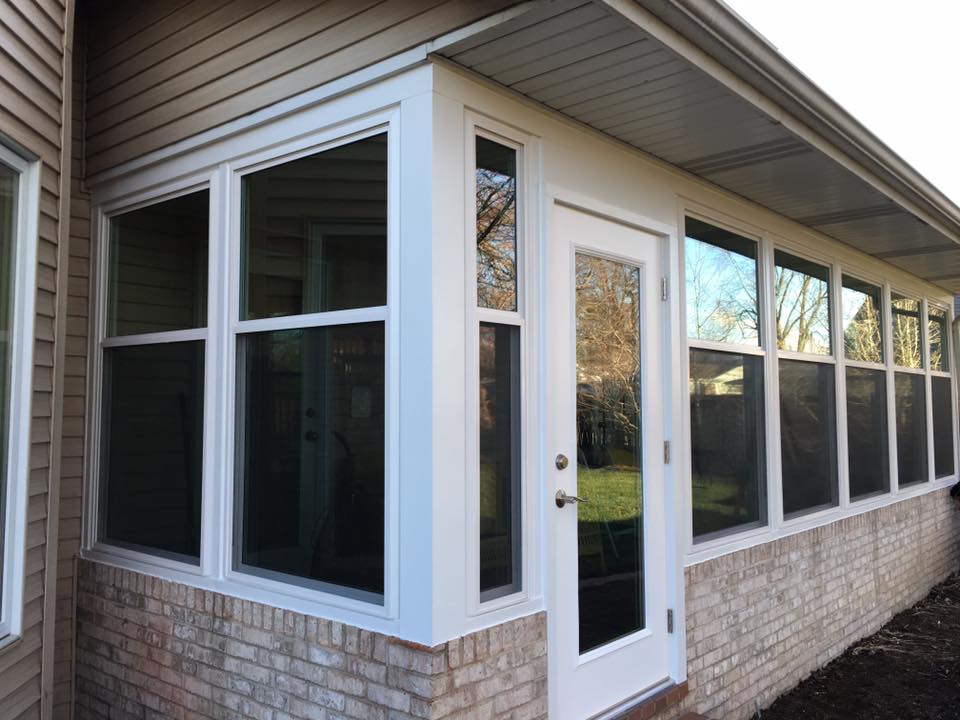 Sunroom with white trim, windows, and door, built onto a brick foundation, attached to a tan-sided house.