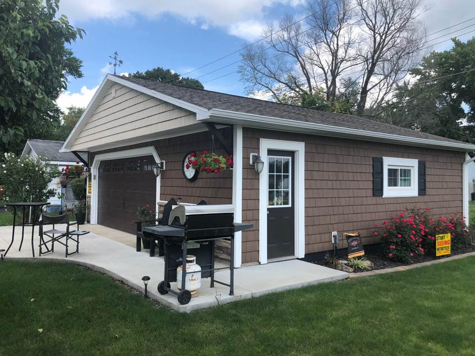Brown shingle-sided garage with a door, window, and grill on a concrete patio, in a yard.