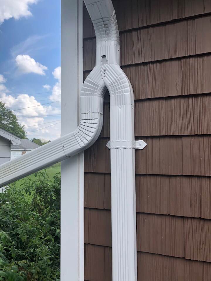 White gutter system on brown shingle siding, with a blue sky and greenery in the background.