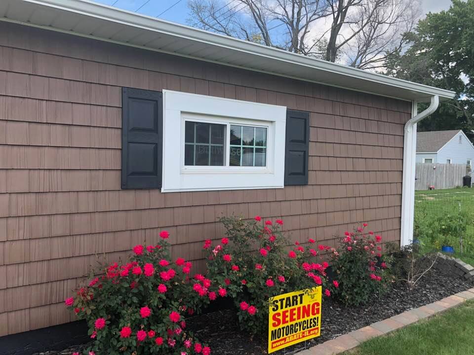 Brown shingle siding with a white-framed window and black shutters, a row of red roses below.