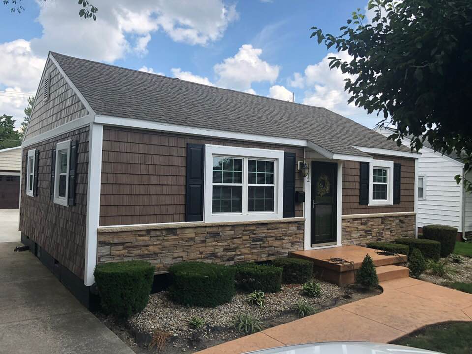 Brown and tan house with stone facade, brown shingles, and dark shutters against a cloudy sky.