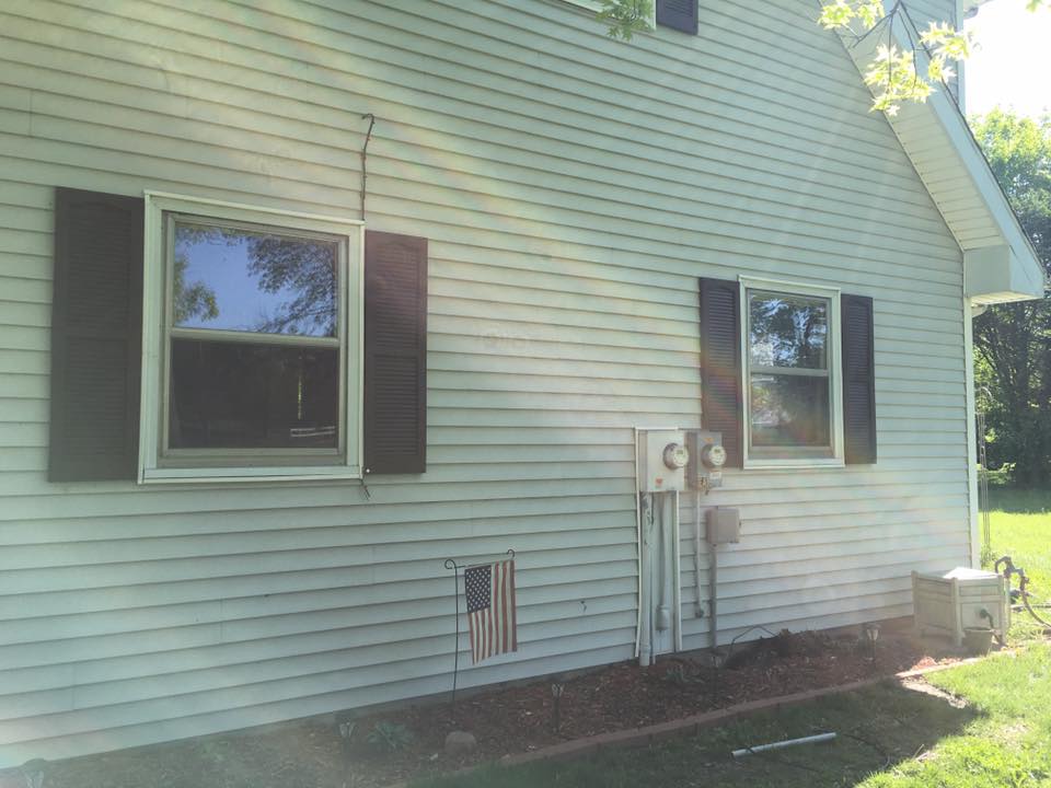Side of a house with two windows, brown shutters, and utility equipment. An American flag is displayed below the windows.
