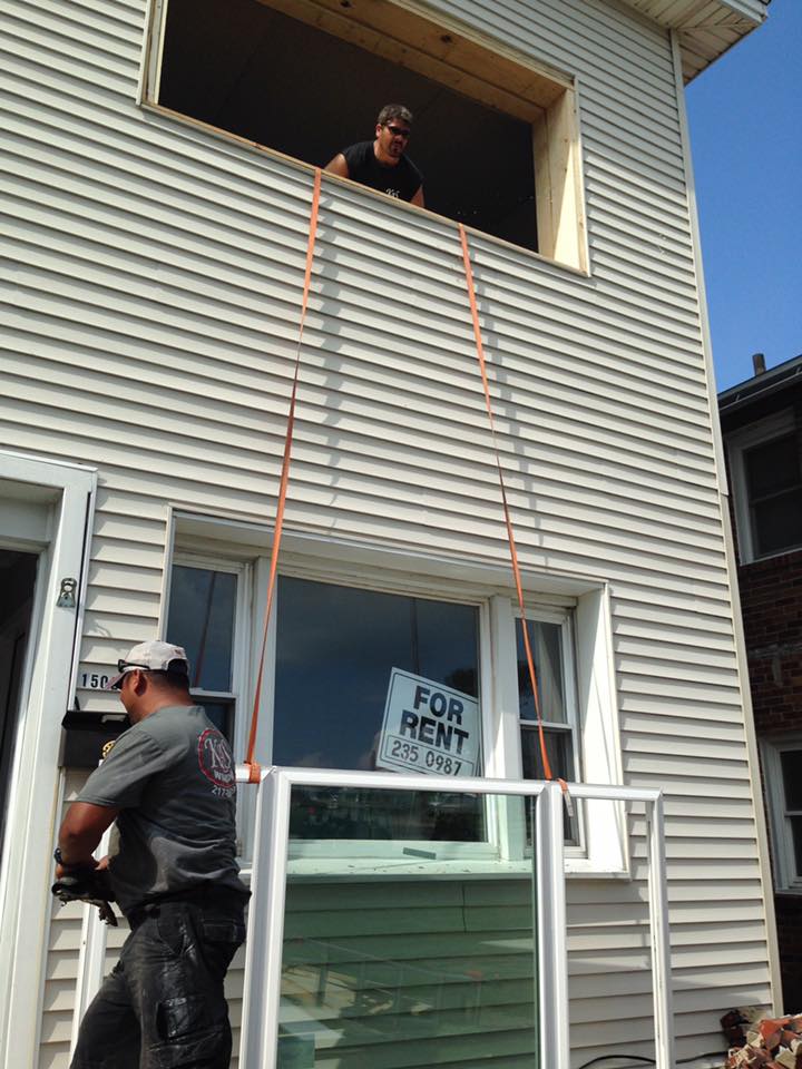 Two workers installing a window: one in the opening above pulling ropes, the other on the ground holding a new window.