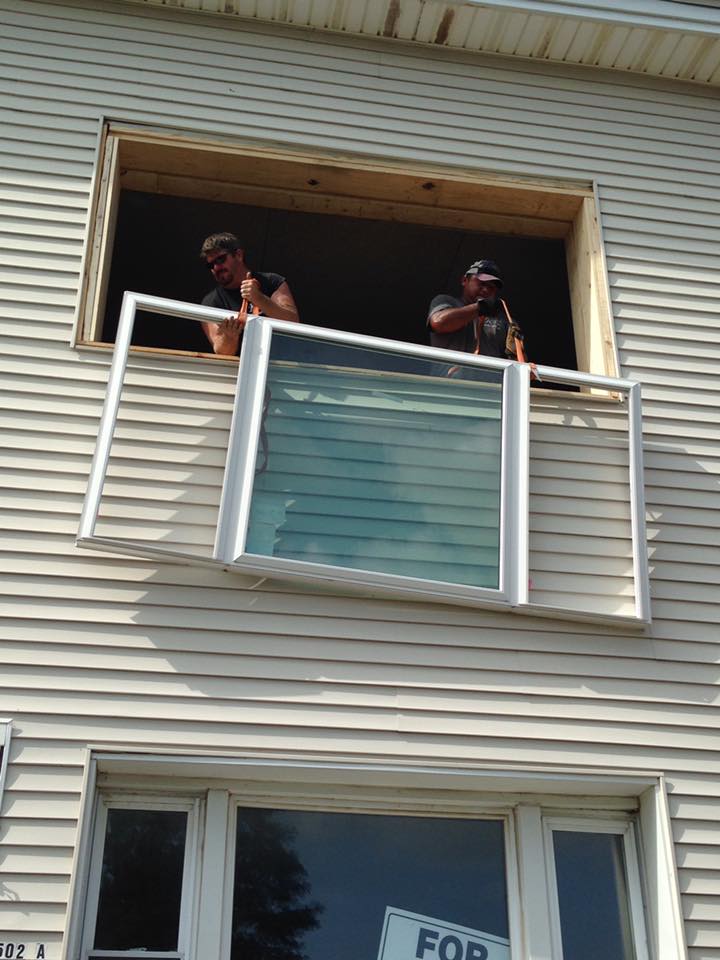 Two men installing a window in a siding-covered house. One man holds a window, the other helps from the opening.