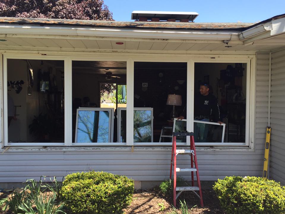 Exterior view of a house, window replacement in progress. Man on ladder, holding a window panel.