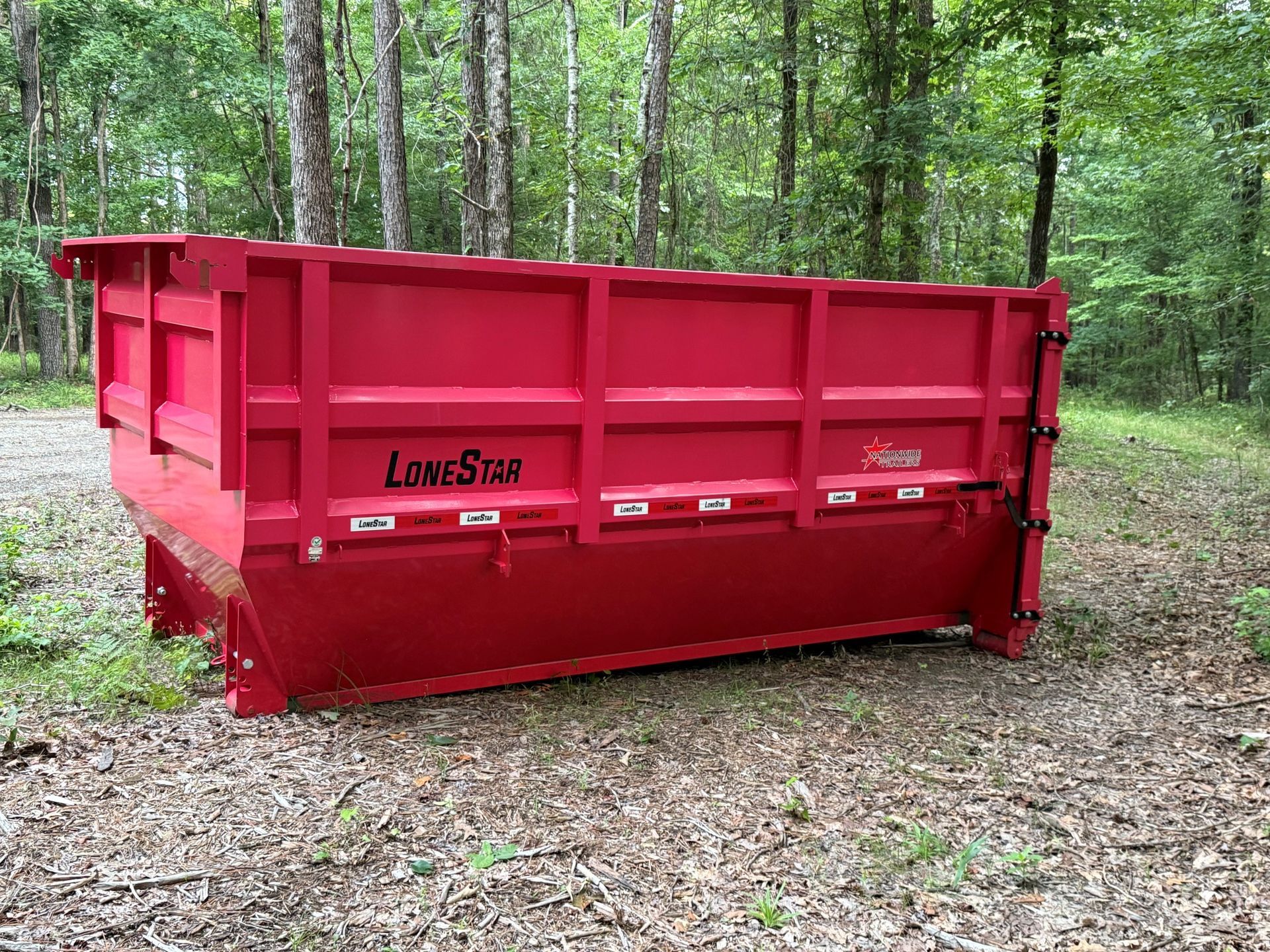 Red roll-off dumpster in a wooded area on dirt ground