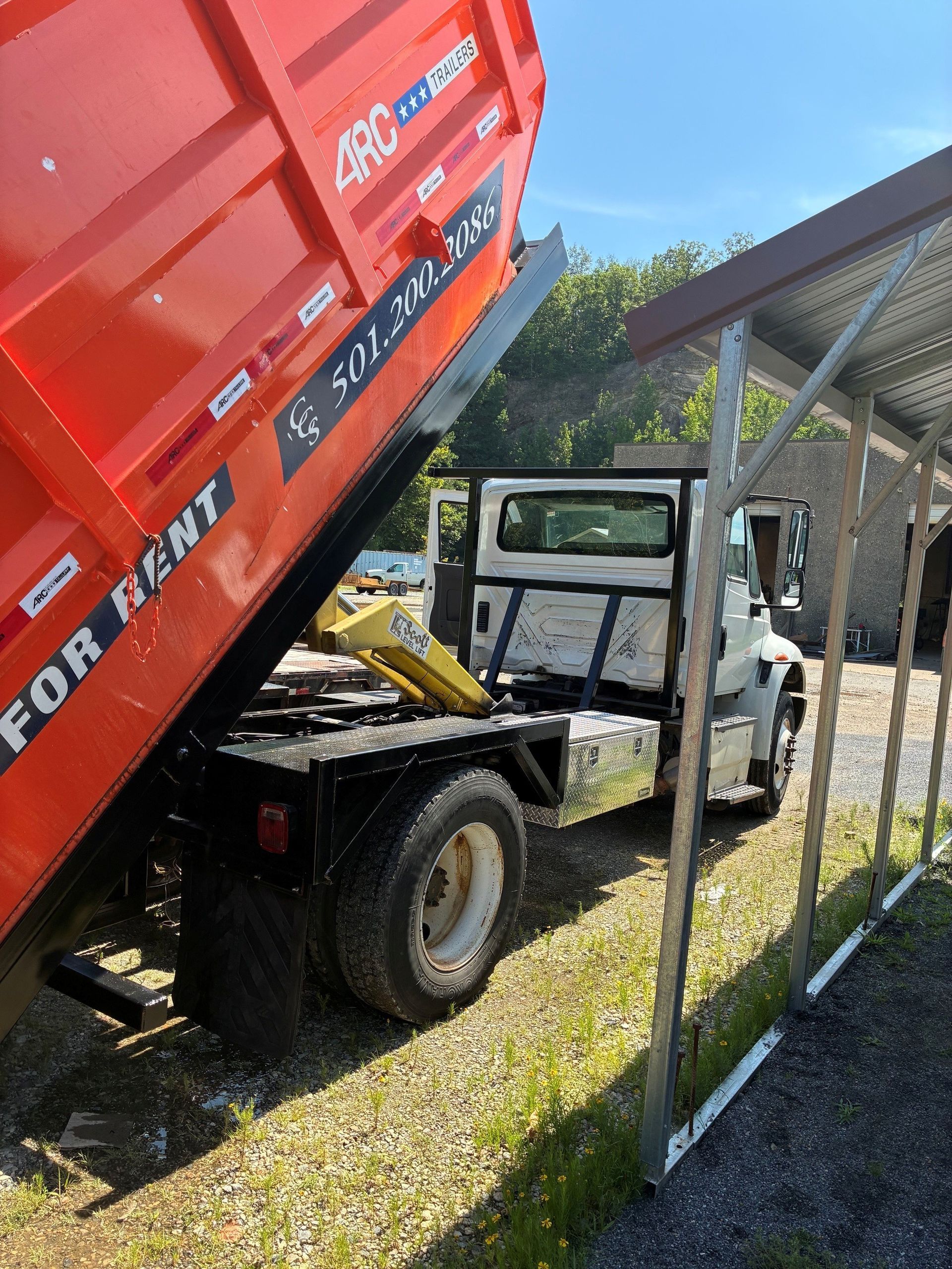 Red dump truck tipping rubble into a small utility truck beside a chain-link fence.