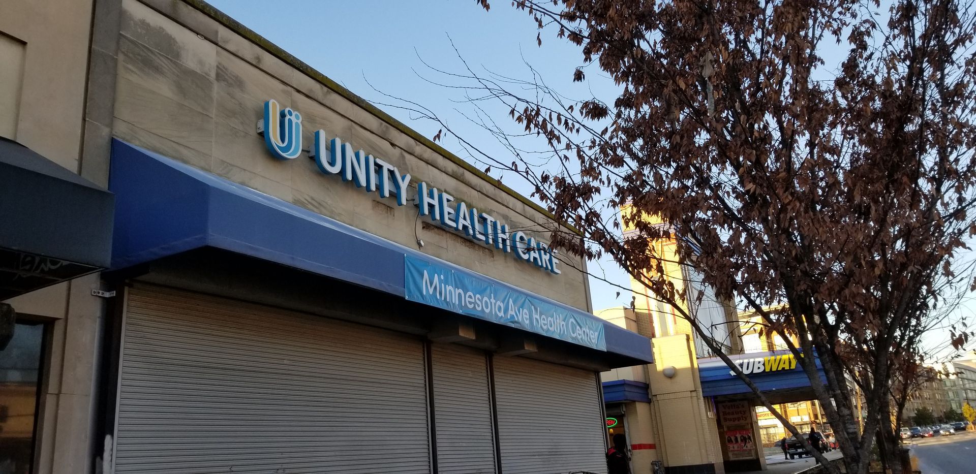 A unity healthcare building with a blue awning and a tree in front of it.