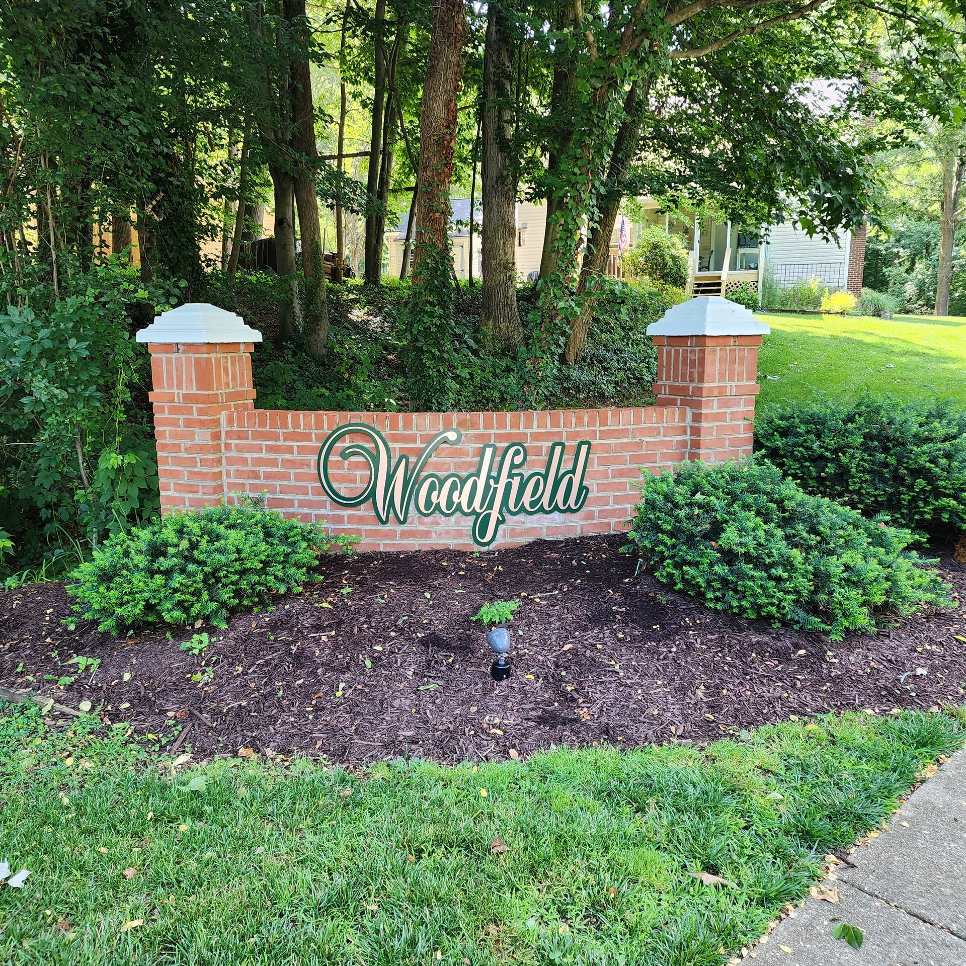 A brick sign for woodfield is surrounded by trees and bushes.