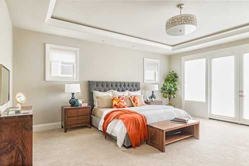 Bedroom with gray headboard, orange throw, and wooden furniture. White walls, doors, and a decorative ceiling light.