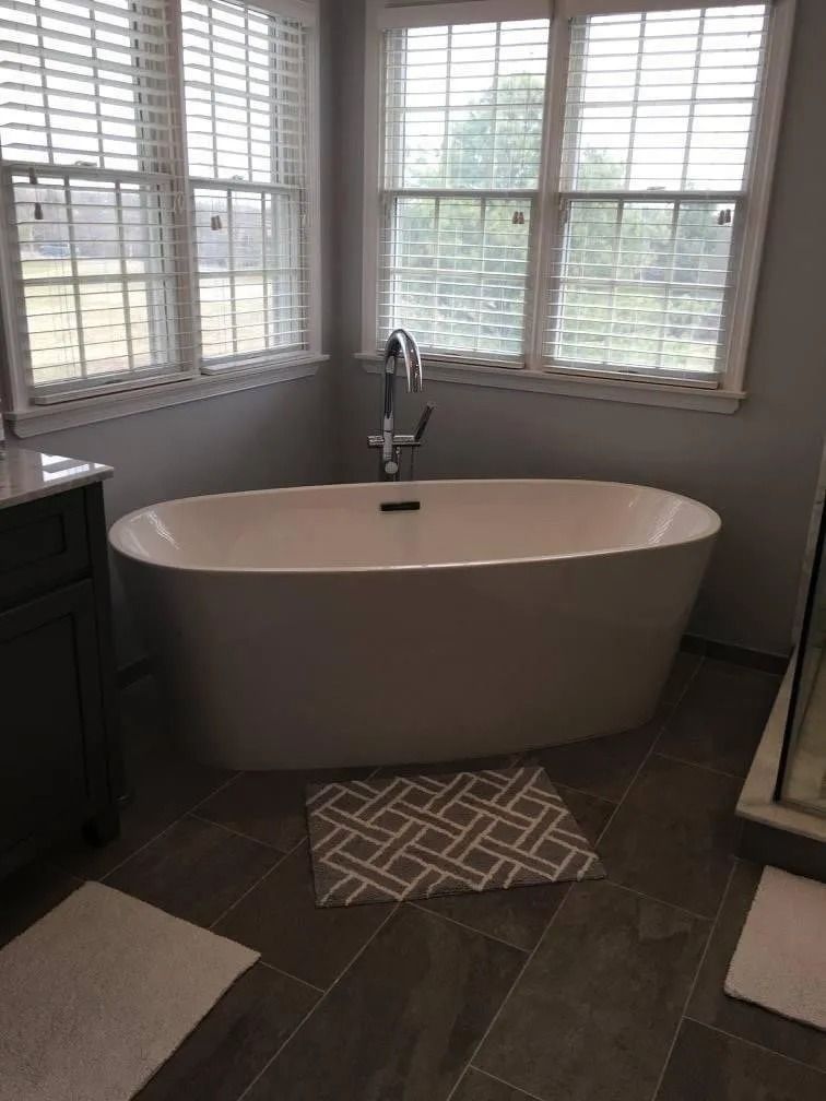 Freestanding white bathtub in a bathroom, near windows with blinds. Gray tiled floor and white bath mats.