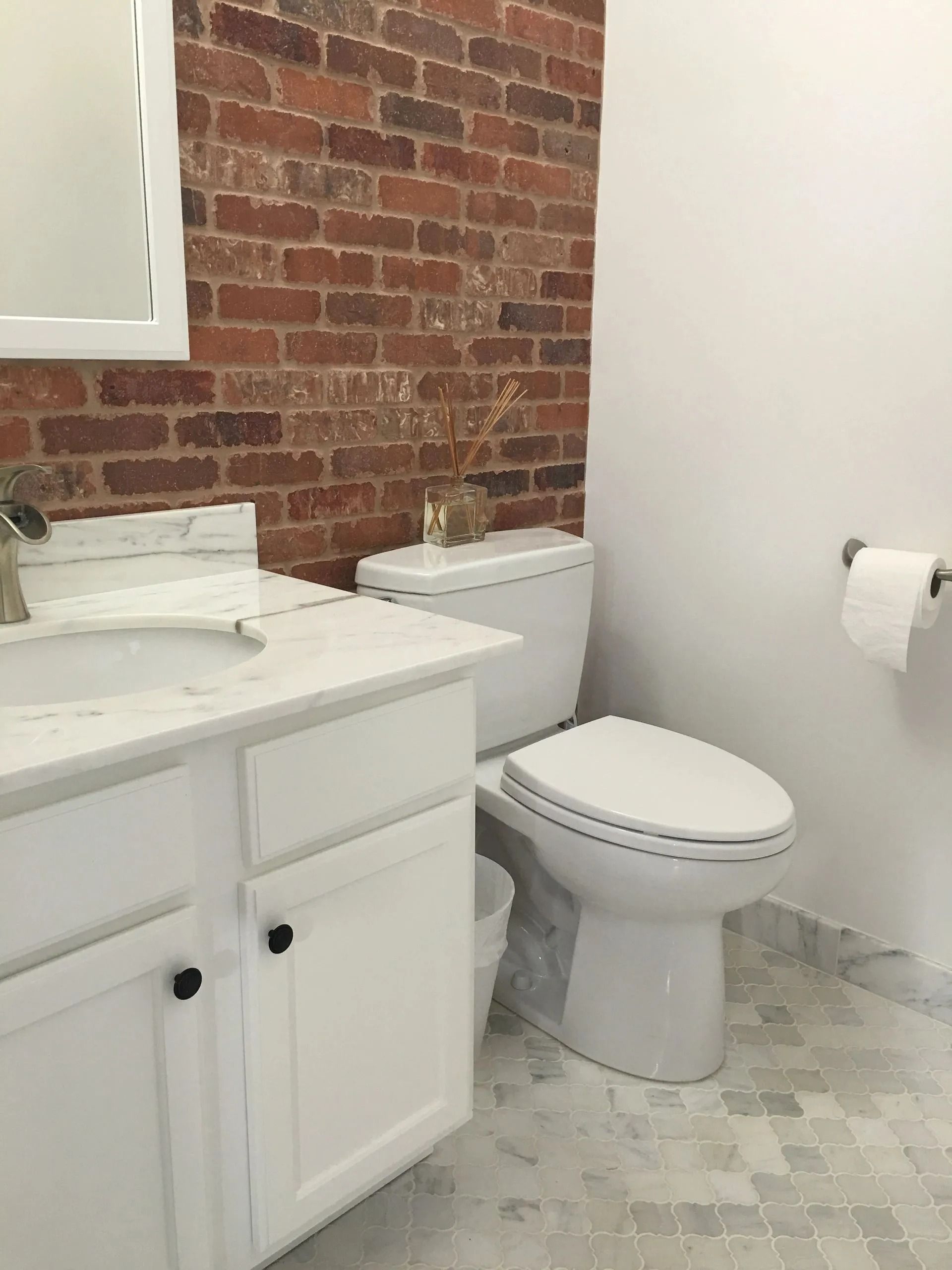 Bathroom with white vanity, toilet, and brick accent wall. Gray and white patterned tile floor.