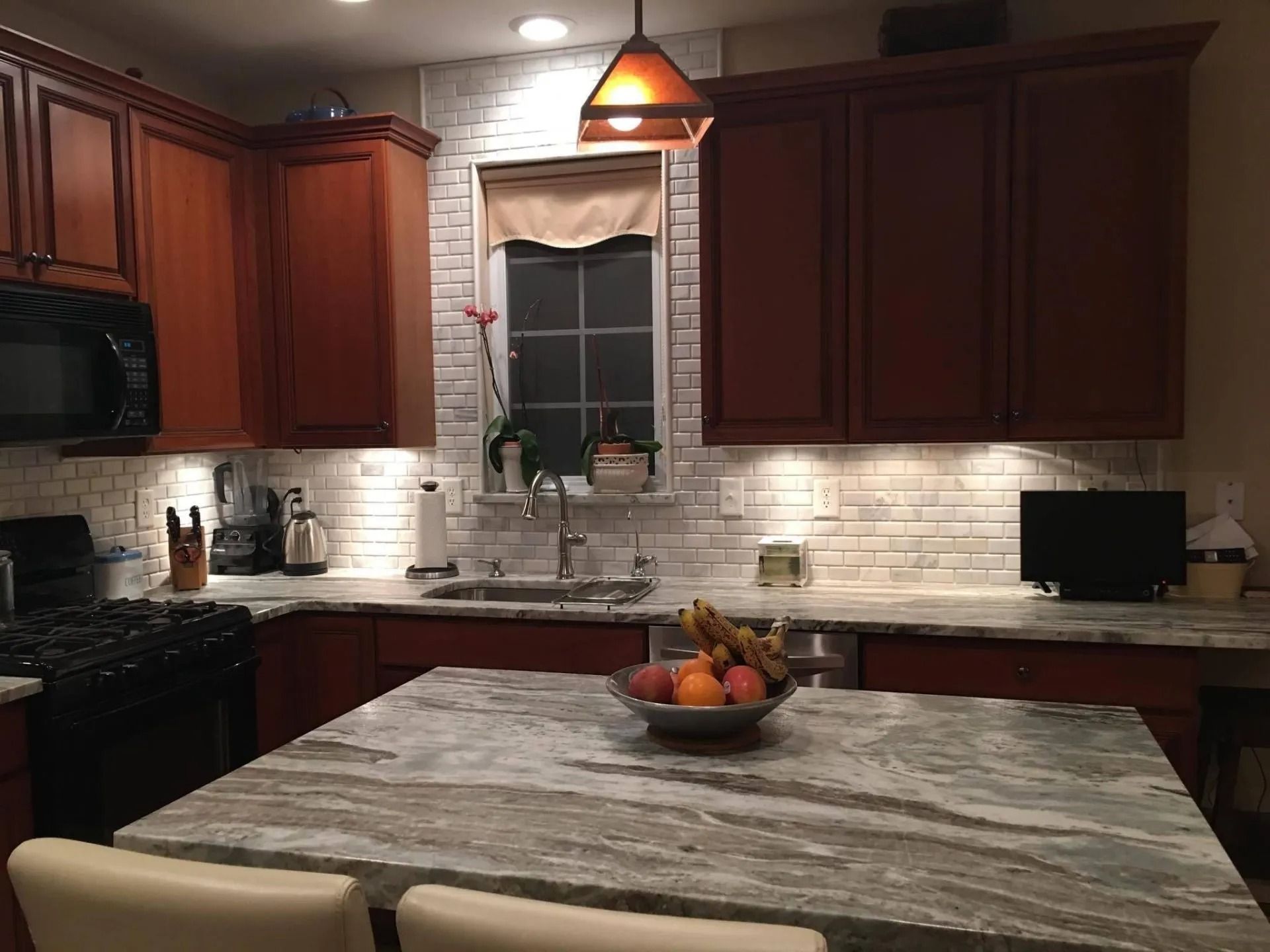 Kitchen with brown cabinets, white backsplash, island with fruit bowl, and a window.