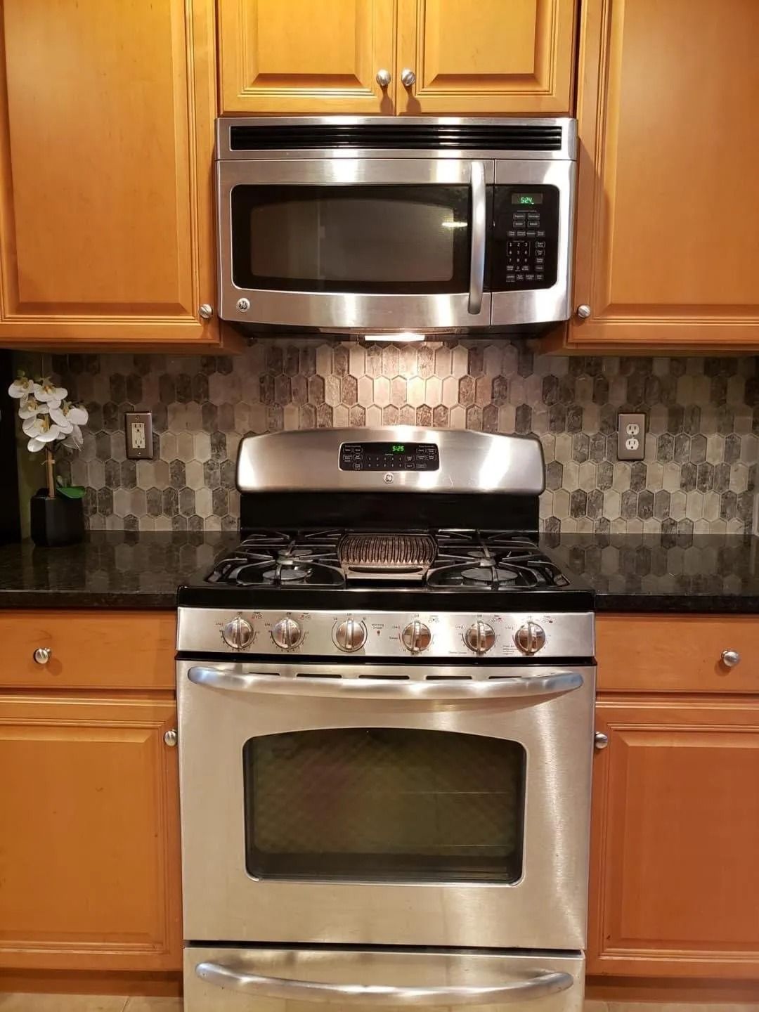 Stainless steel stove and microwave oven in a kitchen with tan cabinets and patterned backsplash.