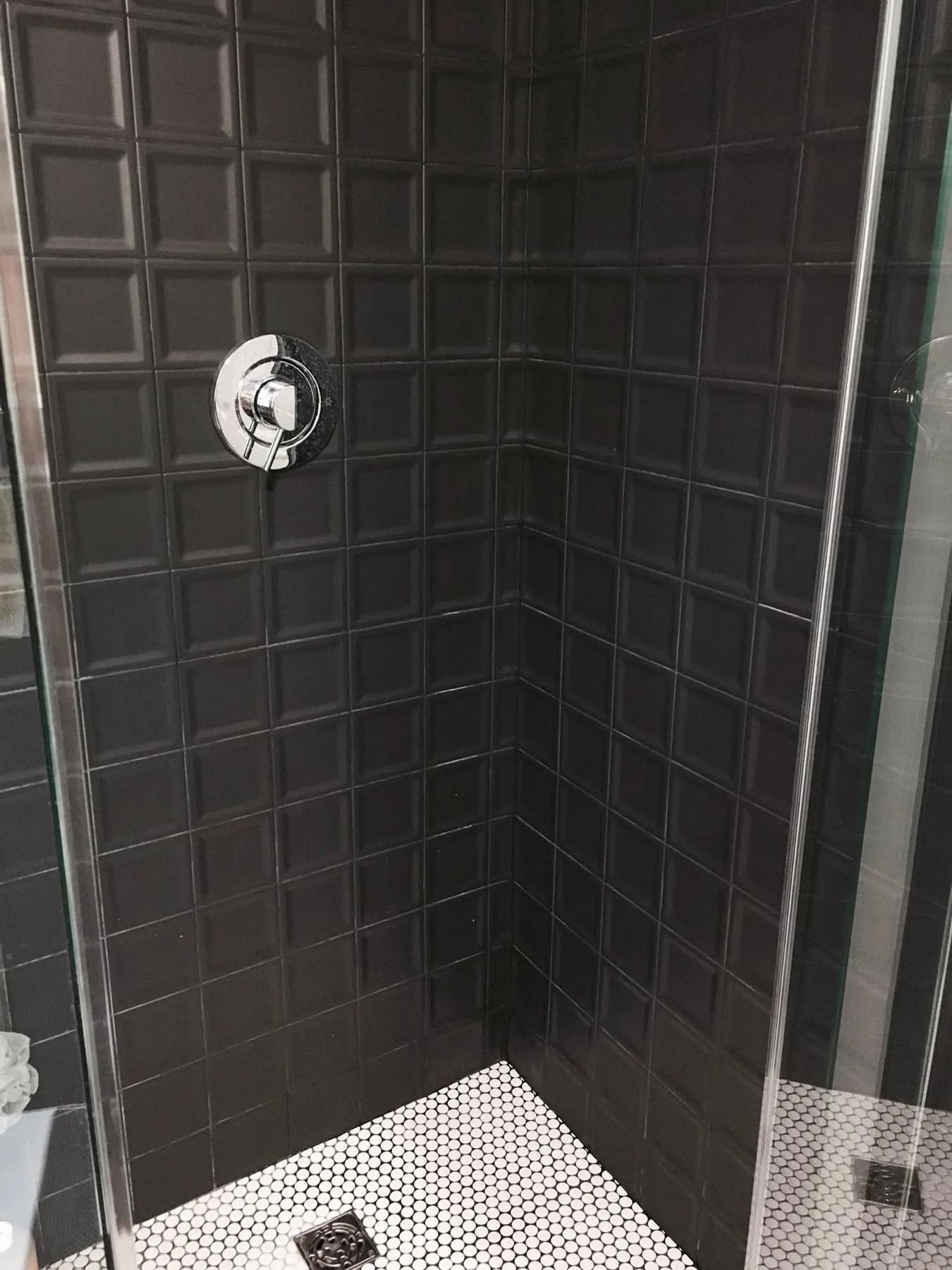 Black tiled shower with a silver faucet, glass door, and white tiled floor.