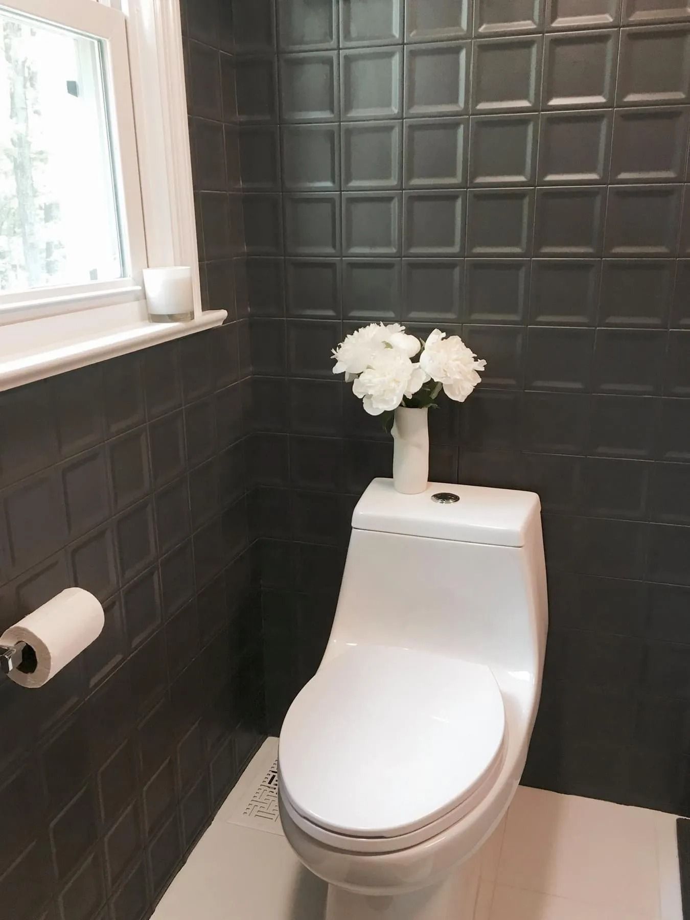 White toilet with white flowers on a black tiled wall in a small bathroom with a window.