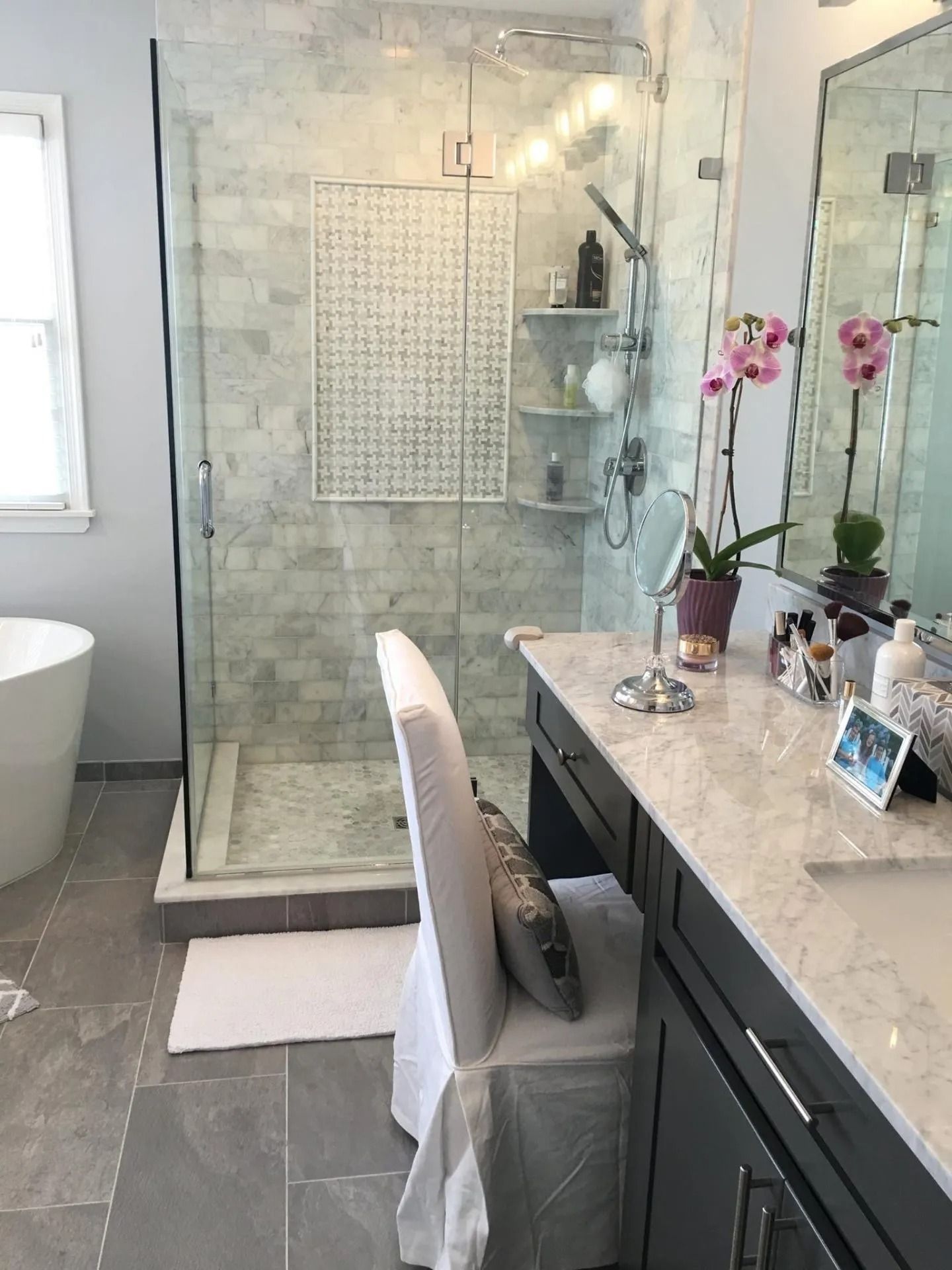 Modern bathroom with a glass shower, vanity, and orchid. Gray tile floor, white chair.