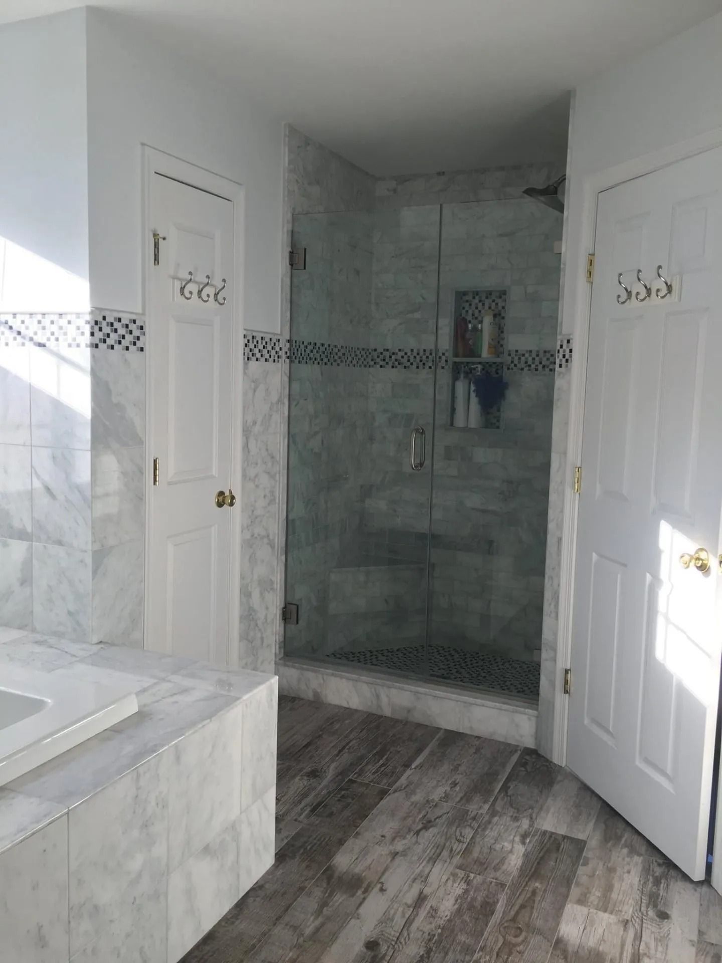 Bathroom with a glass shower, marble walls, white doors, and wood-look flooring.