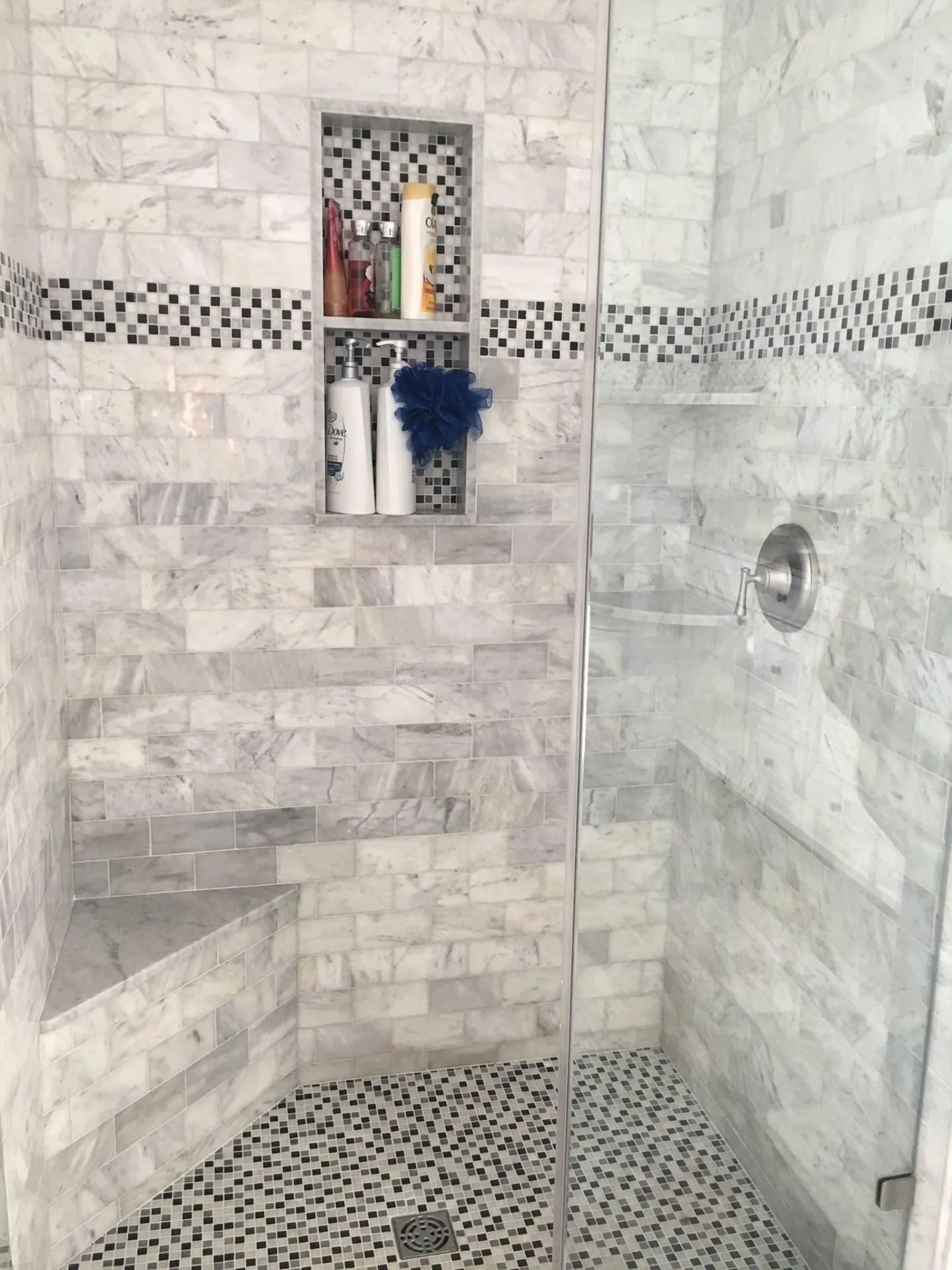 Shower with marble tile walls, pebble floor, built-in shelf with toiletries, and glass door.