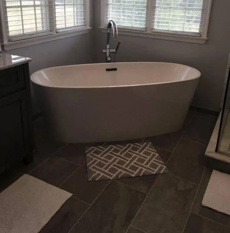 Freestanding oval bathtub in a tiled bathroom with windows and two bath mats