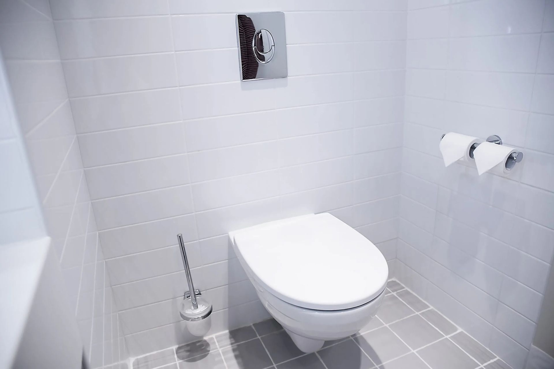 White modern toilet in a small tiled bathroom with a brush and toilet paper.