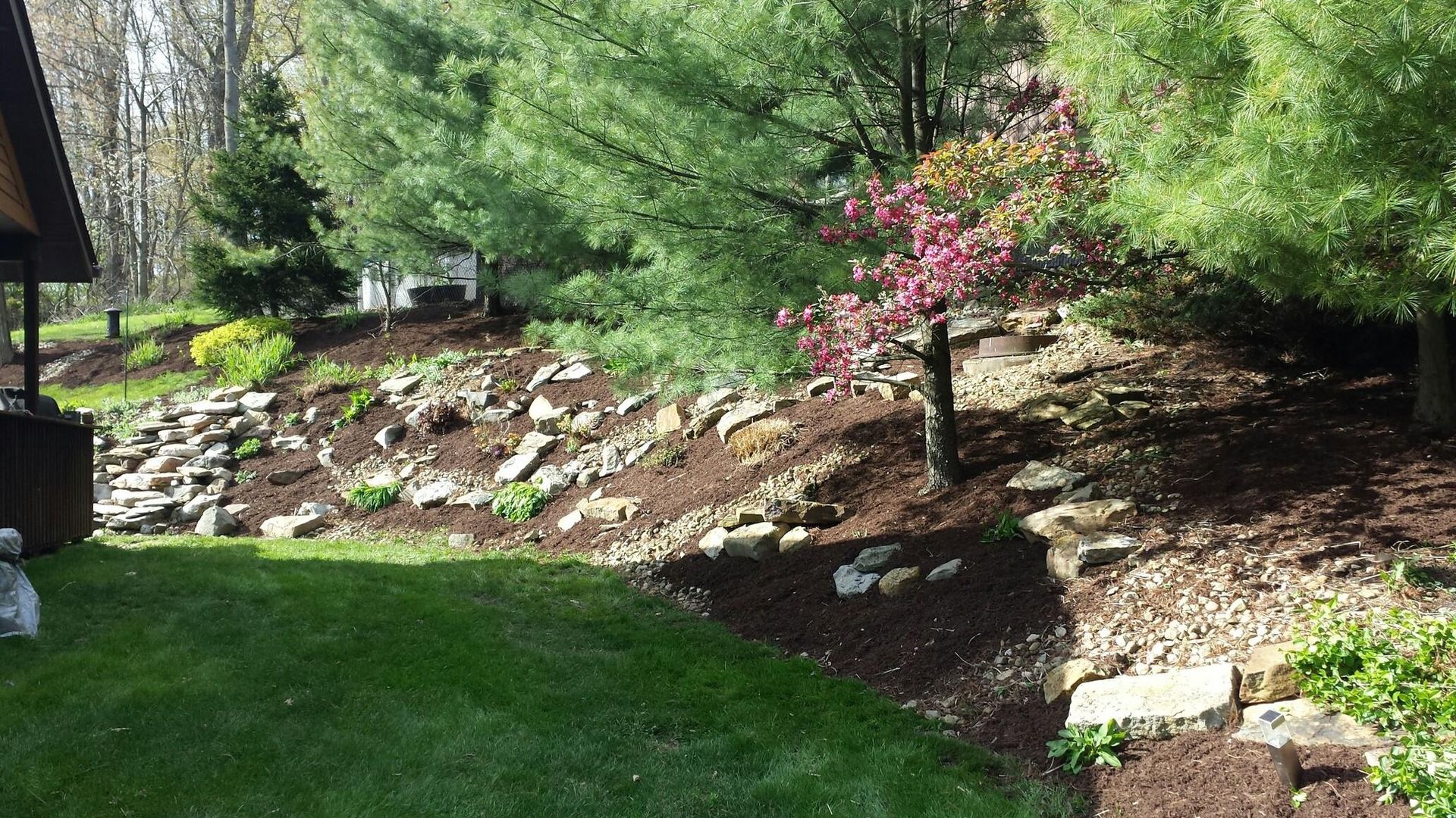 Lush green lawn borders a rocky hillside with trees and reddish mulch. A building's corner is on the left.