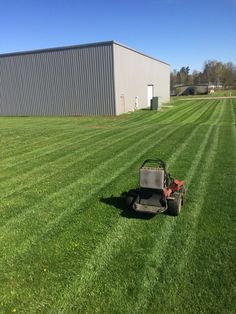 Lawnmower cutting grass in front of a gray industrial building on a sunny day.