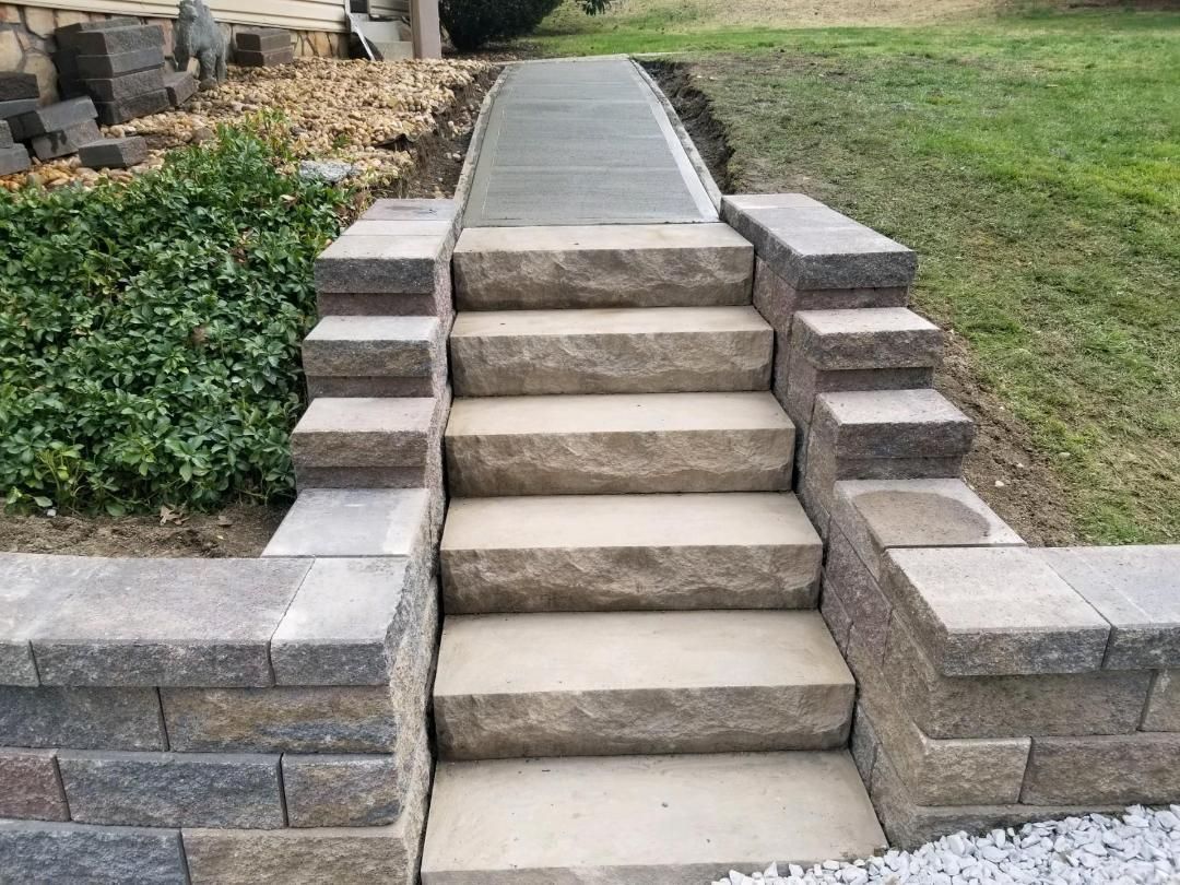 Stone steps leading to a concrete path; bordered by stone retaining walls; green grass in the background.