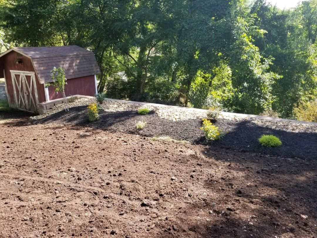 Sloped yard with a small red shed in the background, freshly tilled soil in the foreground, and shrubs planted.
