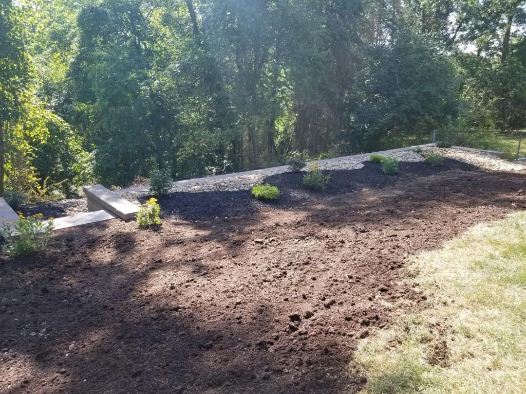 A tiered garden bed with newly planted shrubs, covered in dark mulch, next to a grassy lawn.
