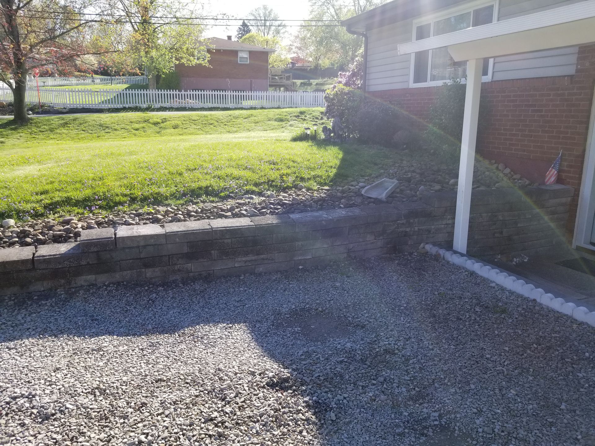 Gravel driveway next to a brick house with a grassy lawn, a retaining wall, and a white fence.