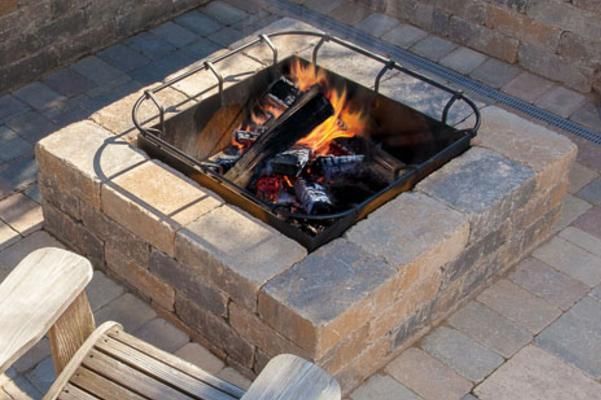 Square brick fire pit with burning logs, metal grate, and patio pavers.
