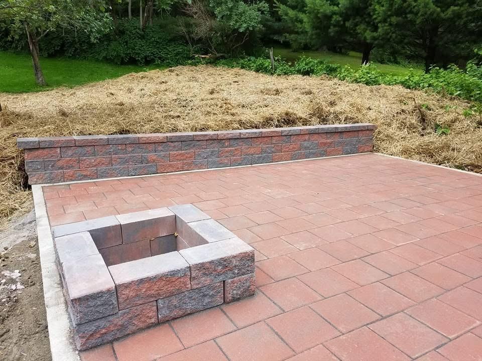 Brick patio with built-in fire pit and retaining wall; surrounded by grass and trees.