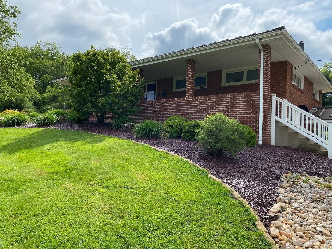 Brick house with white railing and porch, surrounded by green grass, bushes, and trees.