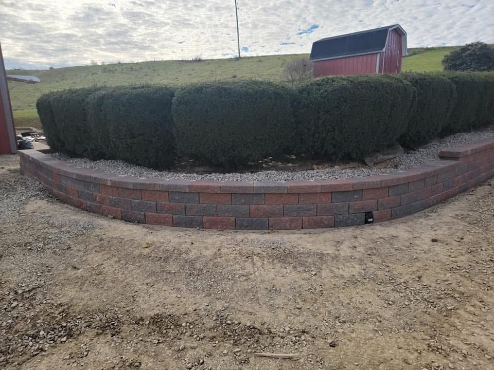 Low brick retaining wall with green hedges on top, against a grassy hill and red barn.