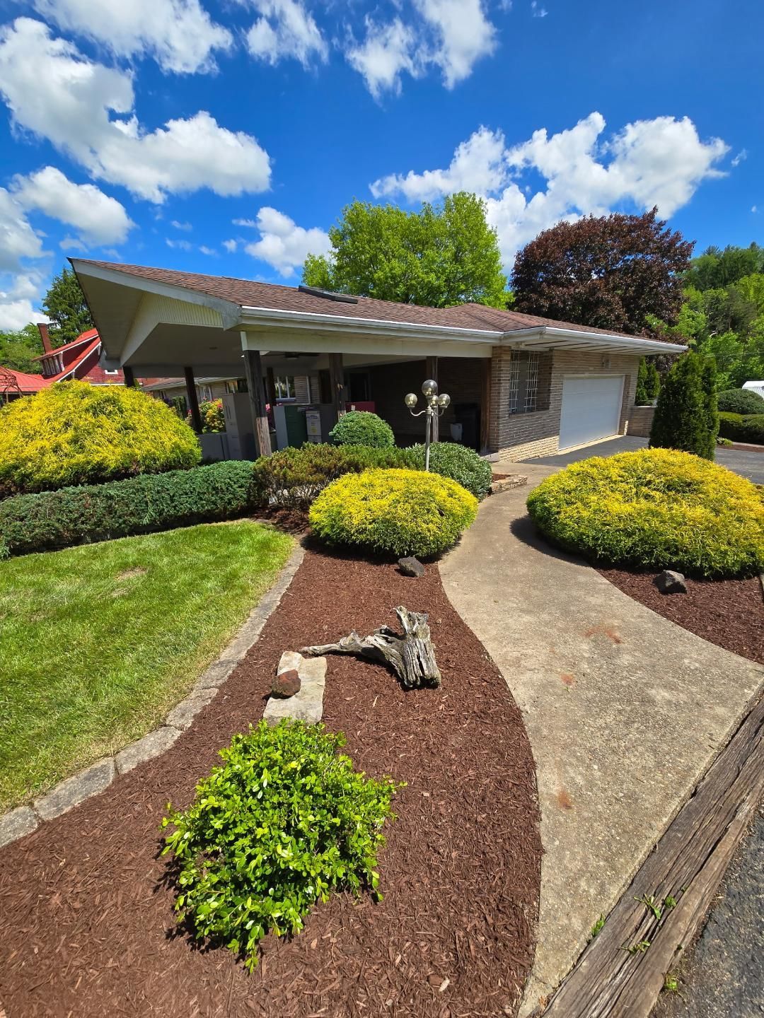 A one-story house with a sloped roof. Landscaping includes green bushes, brown mulch, and a concrete walkway. Blue sky.