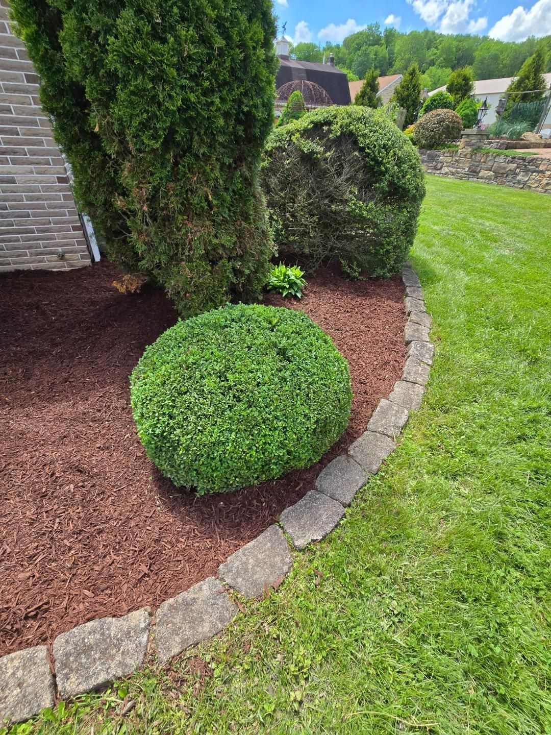 A landscaped yard with a brick building, trimmed bushes, mulch, stone edging, and green grass.