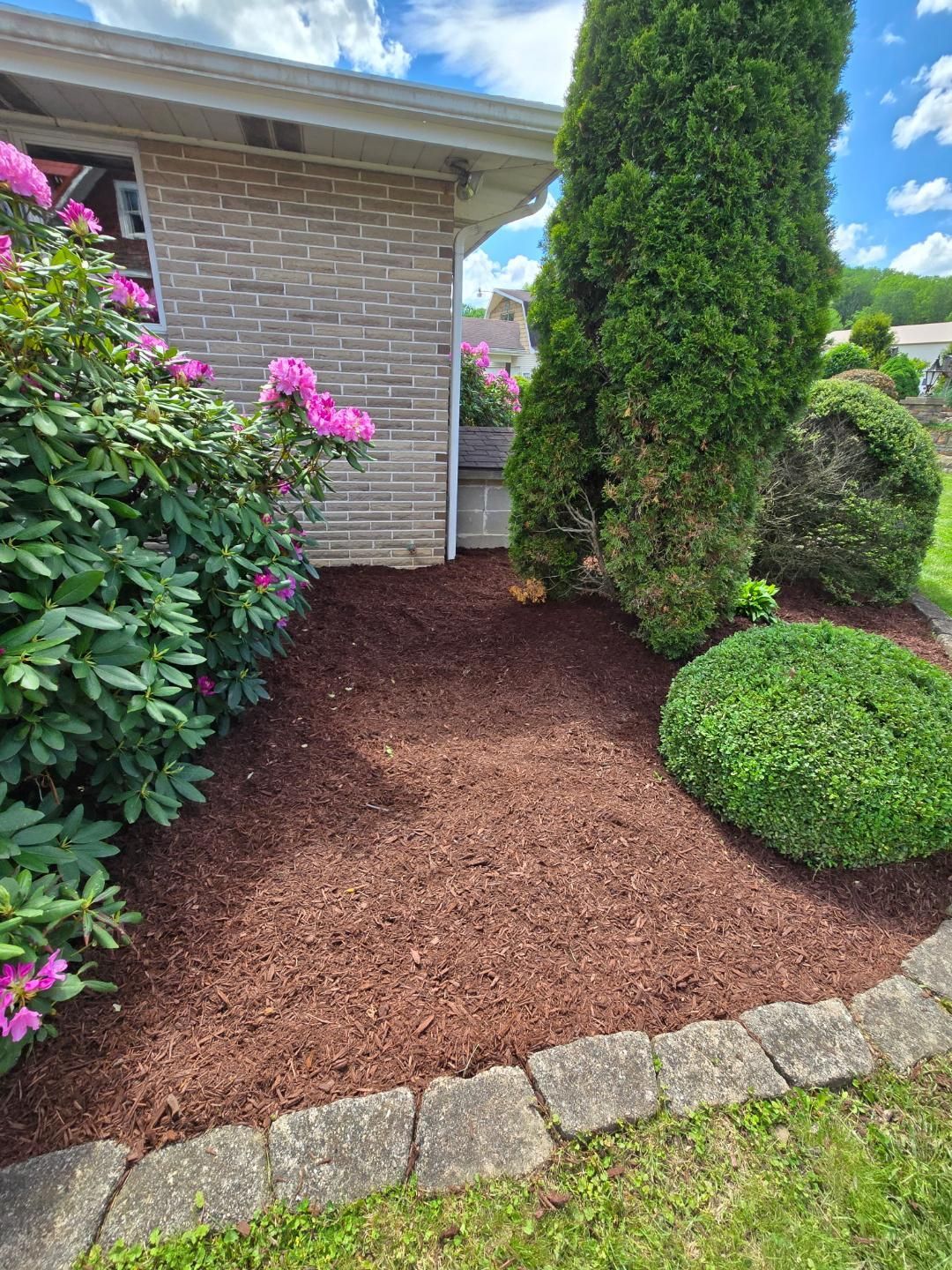 Red mulch garden bed with pink flowers and green bushes, bordered by stone.
