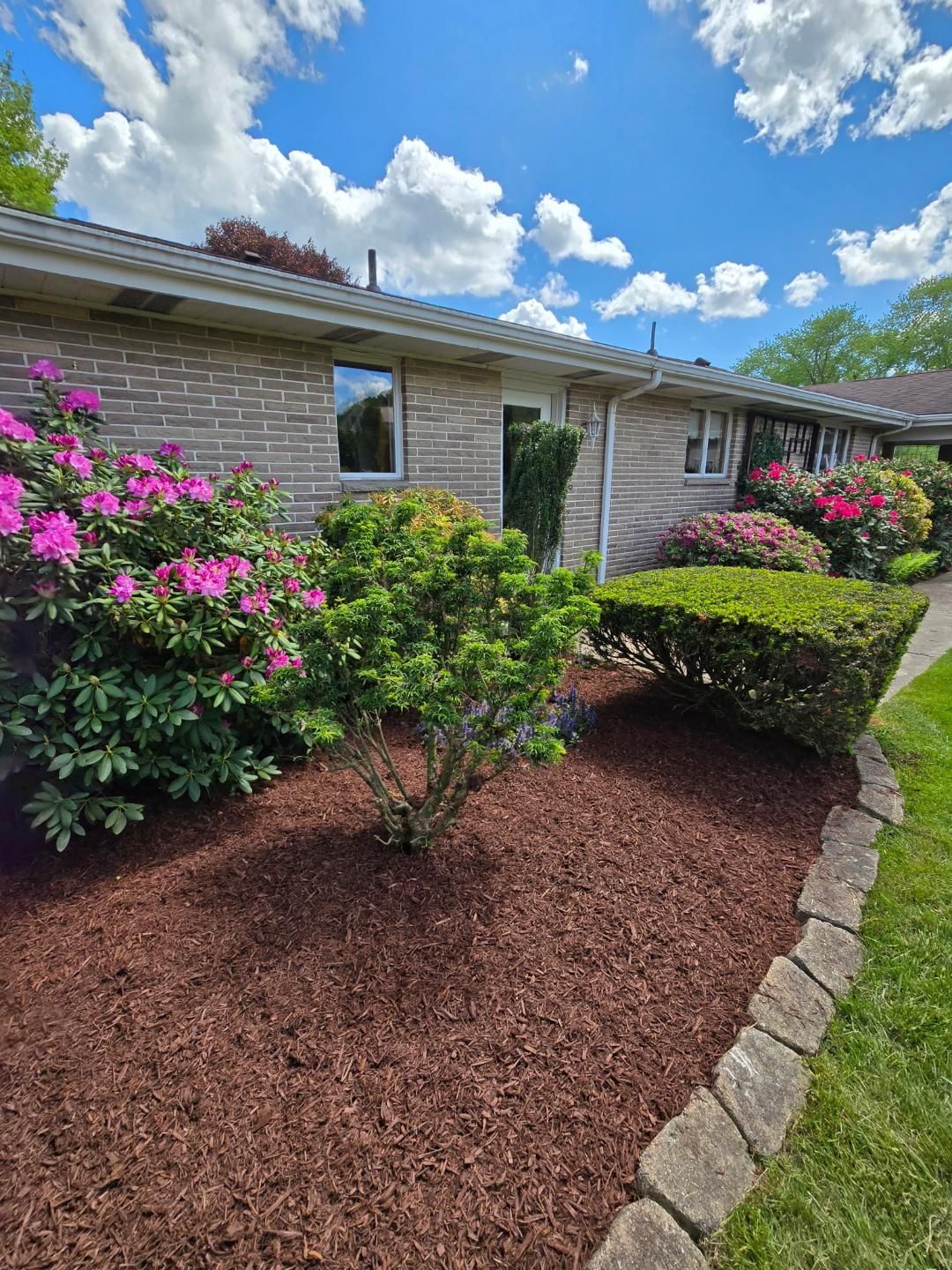 A low-slung brick building with blooming pink flowers, green bushes, and fresh brown mulch under a blue sky.